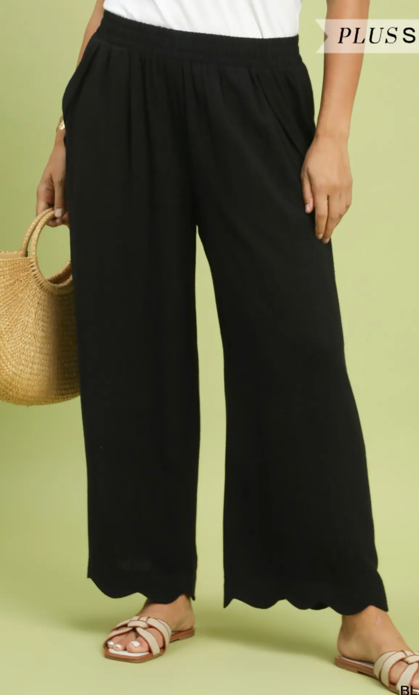 A person wearing loose black wide-leg cropped pants with a scalloped hem, paired with a white top and beige slide sandals, holding a woven straw bag against a light olive green background. The brand name 'PLUSS' is visible in the top right corner.