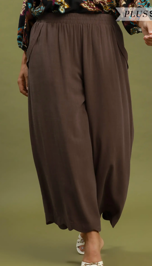 A close-up of a person wearing loose, wide-leg brown pants with an elastic waistband, shown from the torso down against a muted olive-green background.