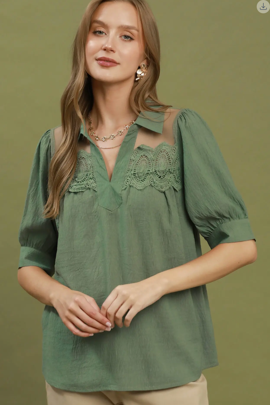 A woman wearing a sage green short-sleeve blouse with lace detailing across the chest, paired with beige pants, against a solid olive green background.